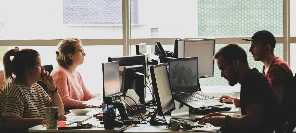 Four colleagues at their desks in a bright open-plan office, two of them talking over the monitors.